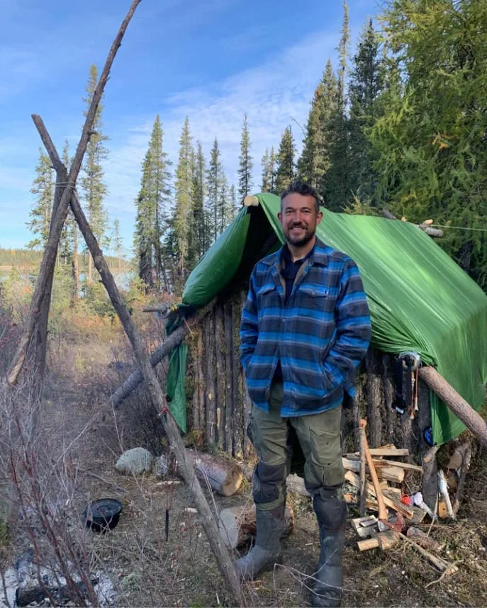 Alone UK survivor stands by his wilderness shelter and campfire.